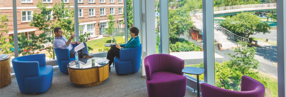 Two faculty members in conversation inside HBS building with view of campus through glass windows